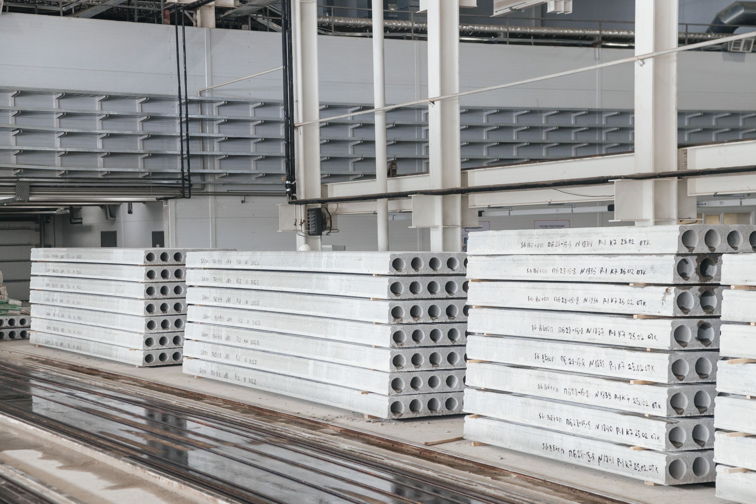 Stacked precast hollowcore slabs with round voids visible on the ends inside a bright production hall, casting beds in the background.