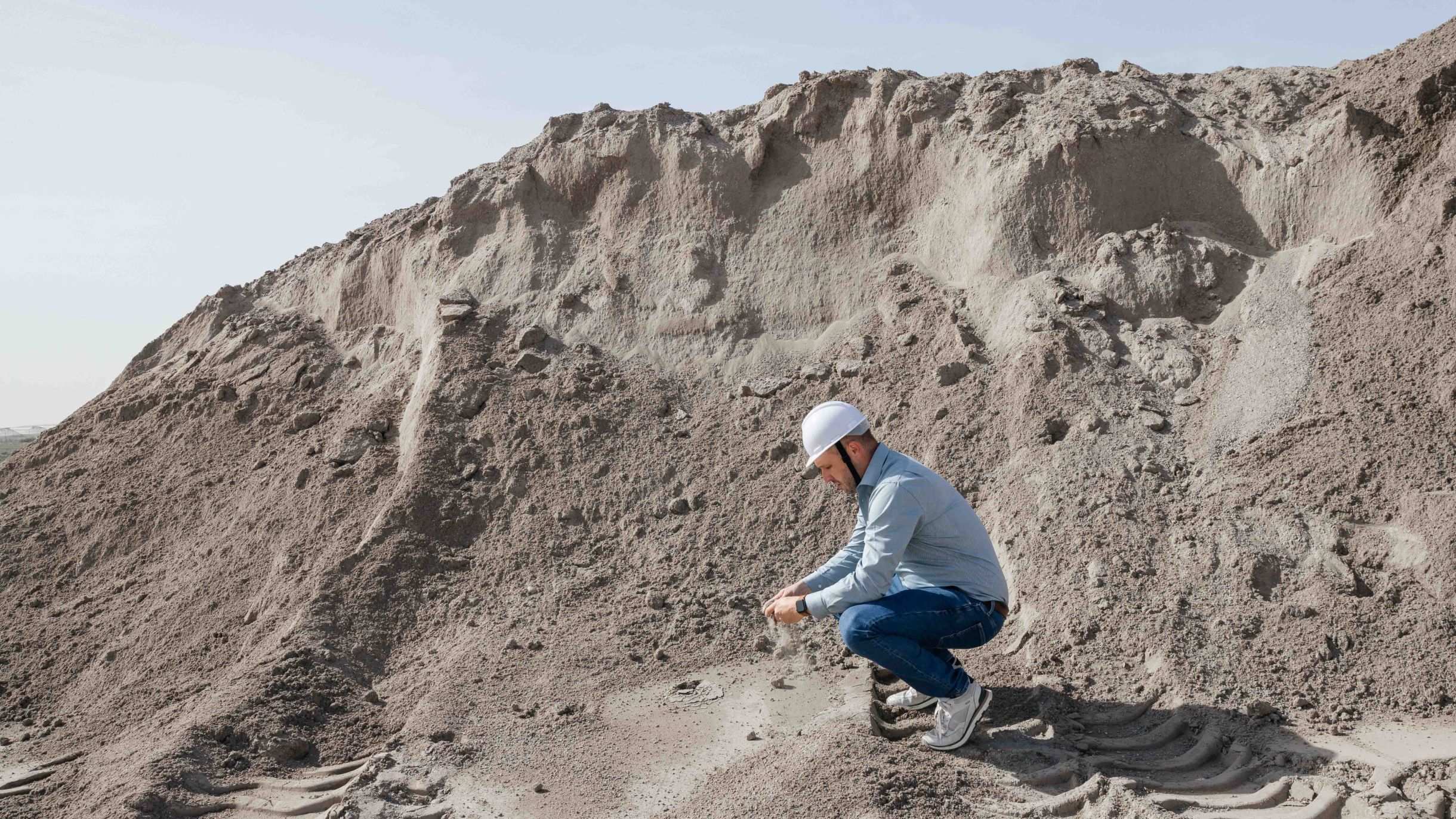 A man wearing a safety helmet is inspecting a large gravel pile in a raw material storage area. He is kneeling down and closely examining the material.