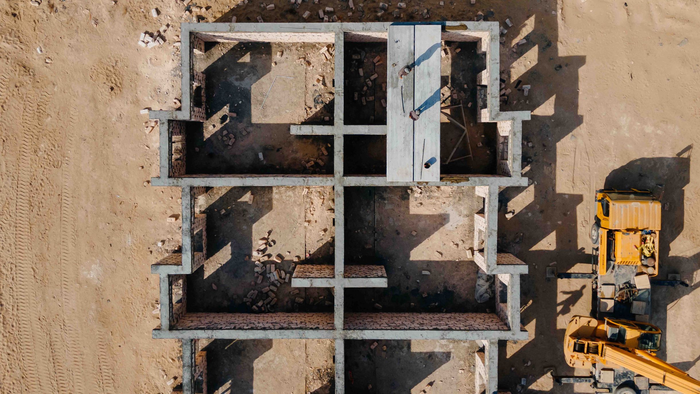 Aerial view of a building shell with a visible floor plan made of precast concrete elements; a yellow crane and a truck are next to it on the construction site.